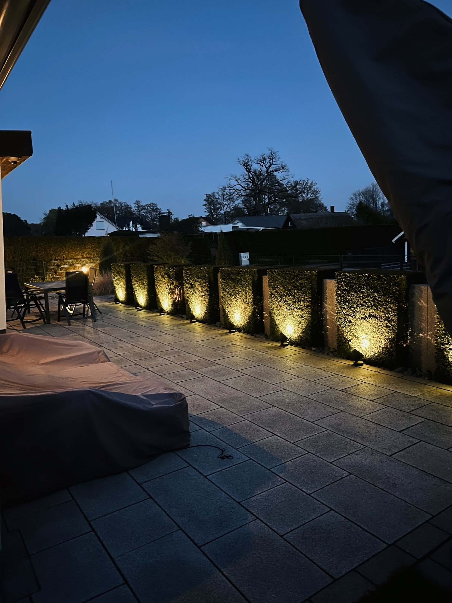 Terrasse abends in der blauen Stunde, beleuchtete Hecke entlang der Granit-Säulen